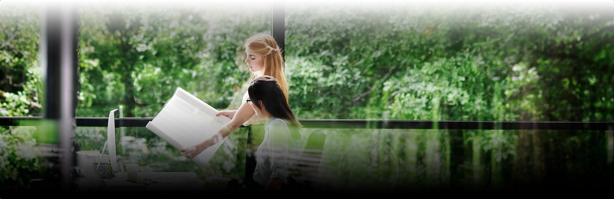 Two young professional women are working at a desk. One of them is holding up a sheet of paper. They are inside a building with glass windows. Trees can be seen in the reflection of the window the photo is taken through.